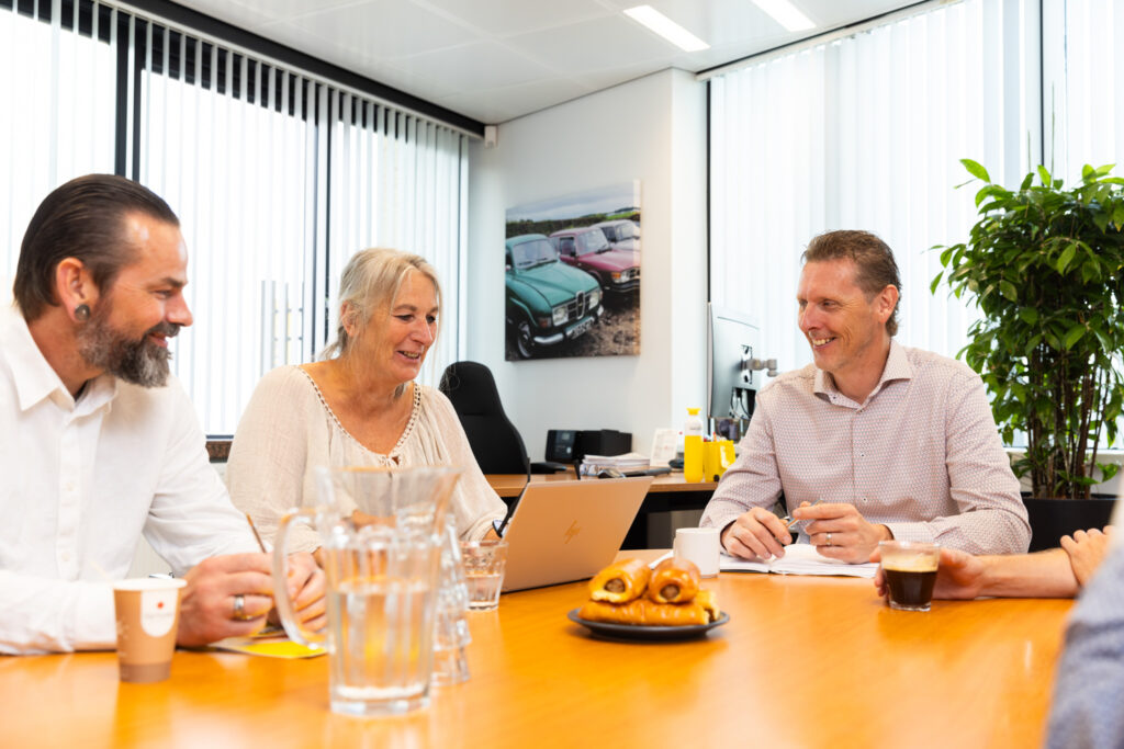 Drie collega’s in overleg aan een vergadertafel met koffie, water en zoetigheden.