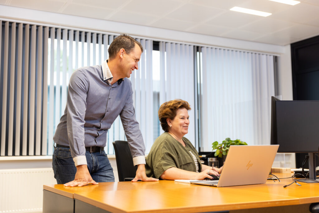 Twee collega’s in gesprek bij een bureau met laptop in een kantooromgeving