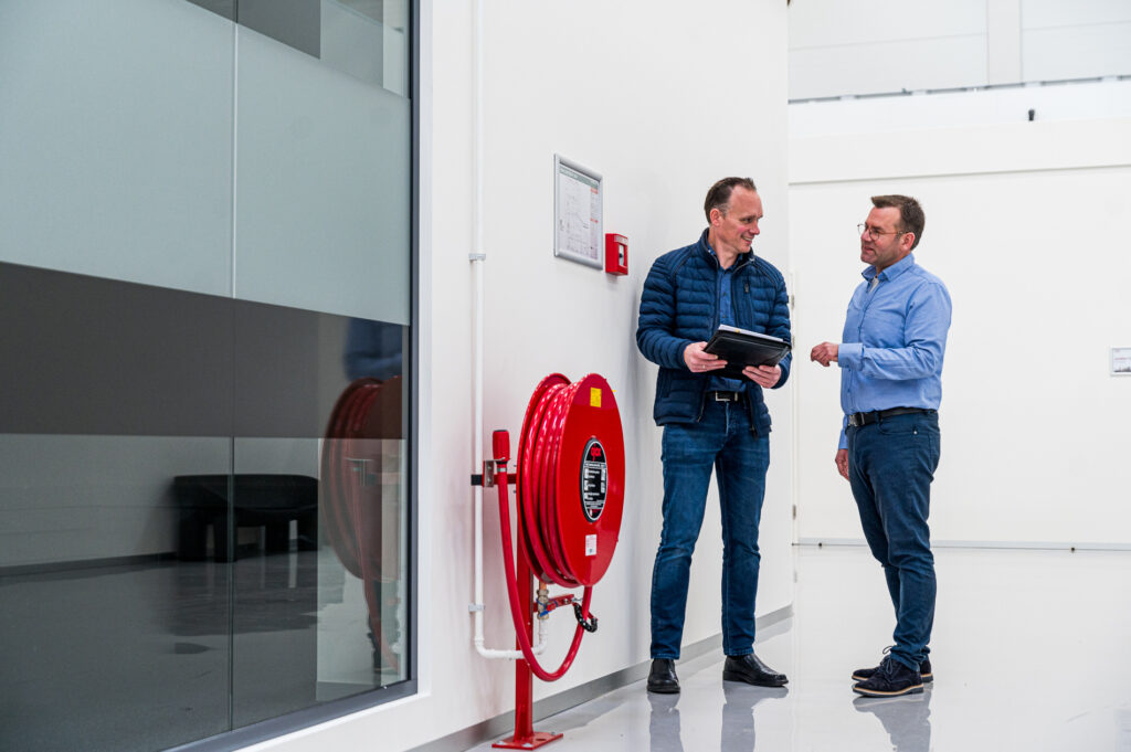 Twee mannen in gesprek in een moderne hal naast een brandslanghaspel, één met tablet in de hand.
