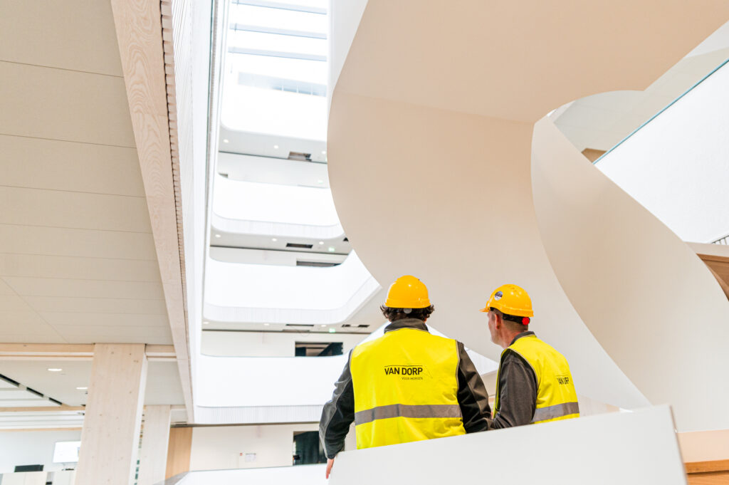Twee medewerkers van Van Dorp met veiligheidshelmen en -hesjes kijken omhoog in een modern atrium.