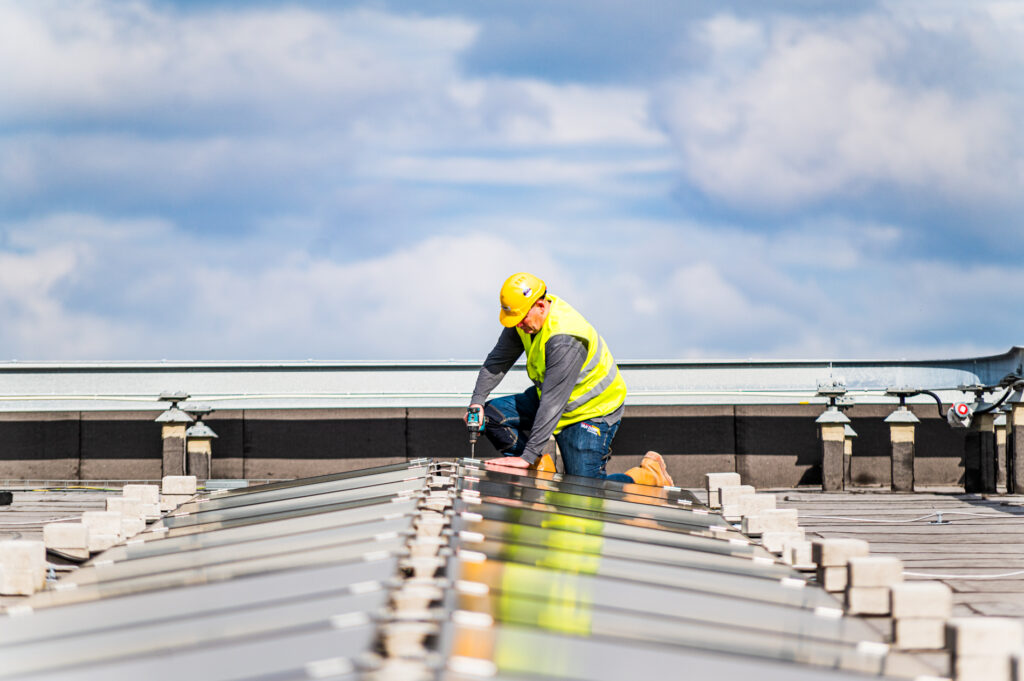 Monteur met veiligheidshelm en geel hesje aan het werk op een dak bij een constructie.