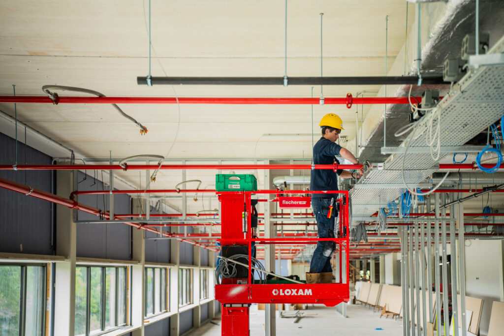 Monteur met veiligheidshelm werkt aan leidingen en bekabeling vanaf een rode hoogwerker in een nieuwbouwpand.