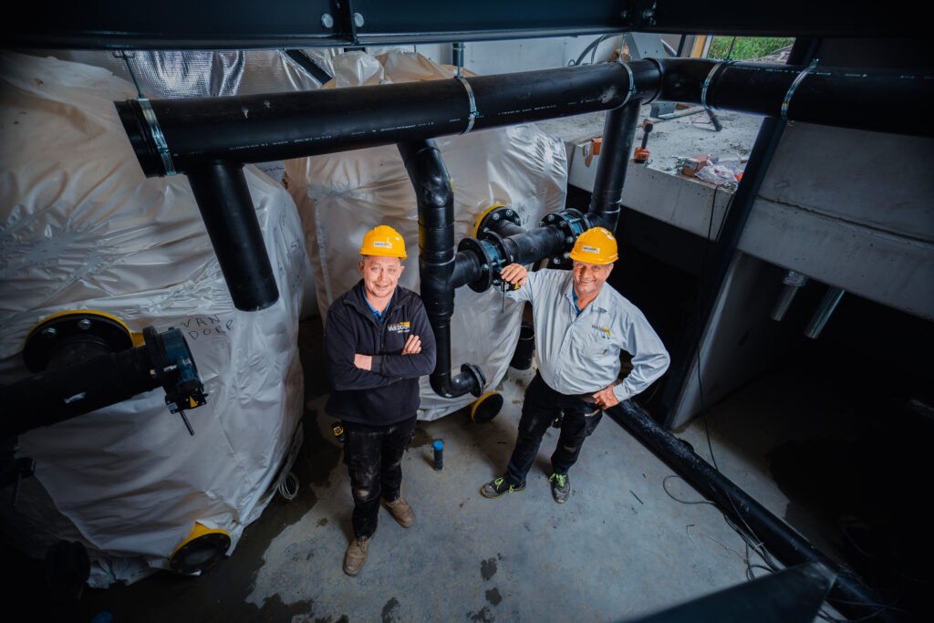 Twee monteurs met veiligheidshelmen in een technische ruimte naast grote installaties en zwarte leidingen