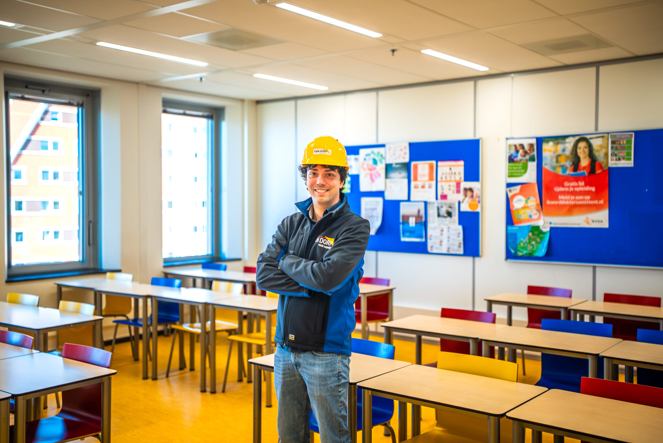 Monteur met veiligheidshelm poseert in een klaslokaal tussen de tafels en stoelen.